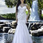 A bride in a long white wedding gown, standing gracefully in front of a small water fountain in an outdoor area, surrounded by trees and plants