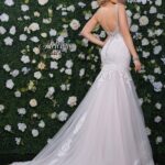 A girl in an ivory white long wedding gown, turned back and striking a pose in front of a wall decorated with white flowers and green plants