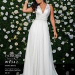 A girl in an ivory white long gown, posing in front of a wall full of white flowers and green plants