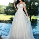 A bride in a long white wedding gown, standing gracefully in front of a small water fountain in an outdoor area, surrounded by trees and plants