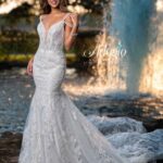 A bride in a long white wedding gown, standing gracefully in front of a small water fountain in an outdoor area, surrounded by trees and plants