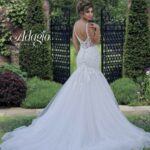 A bride in a long white wedding gown striking a pose by turning back in front of a small gate, with lush green trees and plants climbing a wall behind her, creating a picturesque backdrop