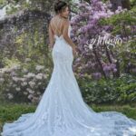 Bride in an ivory white long wedding gown, posing with her back to the camera in front of green plants with purple flowers, on a grassy area