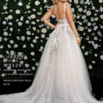 A girl in an ivory white long wedding gown, turned back and striking a pose in front of a wall decorated with white flowers and green plants