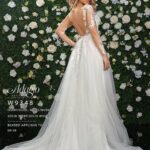 A girl in an ivory white long wedding gown, turned back and striking a pose in front of a wall decorated with white flowers and green plants