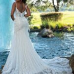 A bride in a long white wedding gown, turned back and striking a pose, standing in front of a small water fountain in an outdoor area surrounded by trees and plants