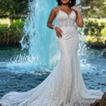 A bride in a long white wedding gown, standing gracefully in front of a small water fountain in an outdoor area, surrounded by trees and plants