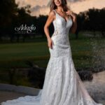 A bride in a long white wedding gown, standing gracefully in an outdoor area, surrounded by trees and plants.