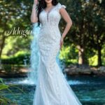 A bride in a long white wedding gown, standing gracefully in front of a small water fountain in an outdoor area, surrounded by trees and plants