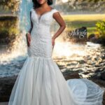 A bride in a long white wedding gown, standing gracefully in front of a small water fountain in an outdoor area, surrounded by trees and plants